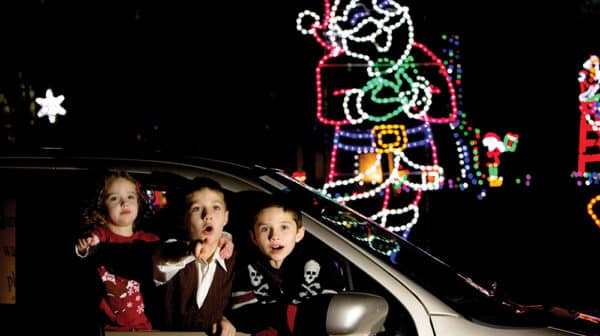 Two young boys and a girl lean out passenger side window of a car, pointing with surprised looks on their faces. Lighted Santa display at the Santa Claus Land of Lights is visible in dark blackground