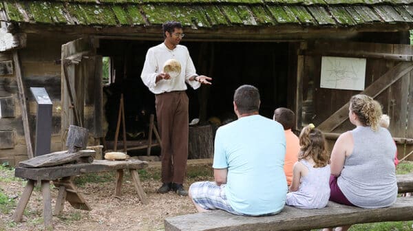 Costumed interpreter at the Living Historical Farm at Lincoln Boyhood National Memorial performs a demonstration in front of a family sitting on wood benches