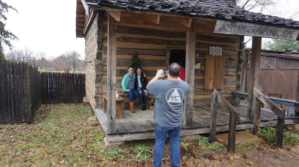 A man takes a photo of two people sitting on a bench on the porch of a log cabin