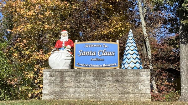Welcome to Santa Claus, Indiana, sign features a blue sign with Santa statue on the left and tree statue to the right, all displayed atop a stone wall; with fall-colored trees in the background