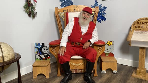 Santa Claus sits in a wooden chair with red accents and Christmas decorations on the walls behind him