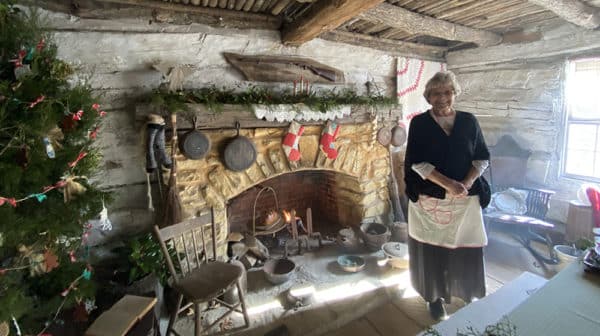 Woman is dressed in black pioneer dress with white apron and stands in front of a stone fireplace next to a Christmas tree in a cabin decorated for the season
