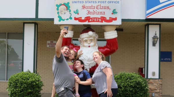 A family of four takes a selfie in front of a Santa statue that stands in front of the Santa Claus Post Office
