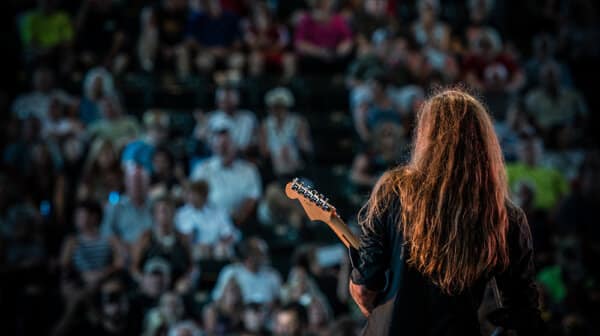 Guitarist with long hair is shown from the back on the stage at Lincoln Amphitheatre as he performs for a crowd