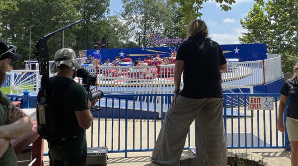 Cameraman and photographer shooting destination video at Holiday World stand with backs to camera with Firecracker ride in the background