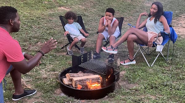 Dad kneels by campfire tells story to wife and two kids sitting in camp chairs at Sun Outdoors Lake Rudolph during destination video shoot
